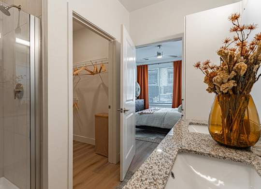 A bathroom with a walk-in shower, a bedroom, and a vase of dried flowers on the counter.
