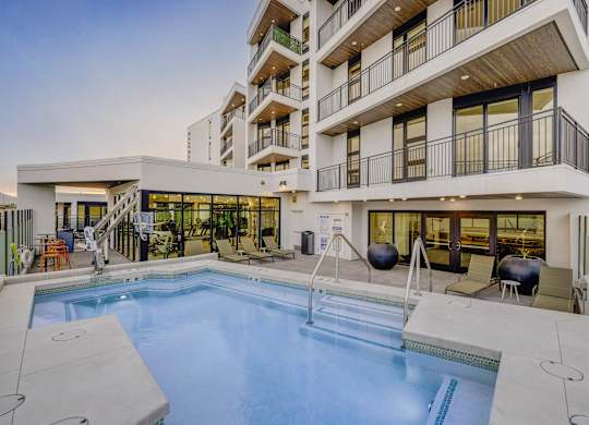 A large swimming pool is surrounded by a patio area with chairs and a hot tub.
