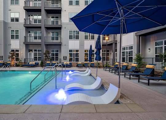 a swimming pool with a blue umbrella and some chairs in front of an apartment building