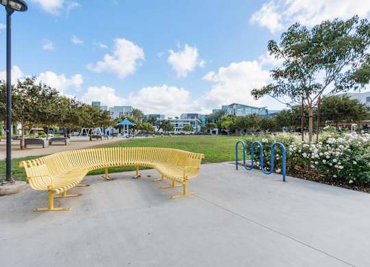 A yellow bench sits in a park with a blue pole behind it.