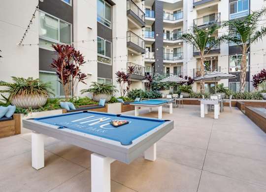 A pool table with a blue cloth and white legs is in the middle of a courtyard with plants and a building in the background.
