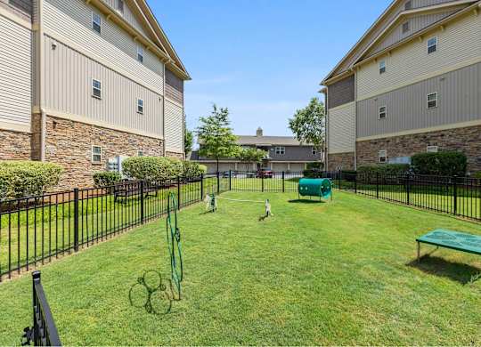 A black fence surrounds a green lawn with a tennis court and a bench.