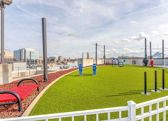 A white fence surrounds a green area with a red bench and blue trash cans.