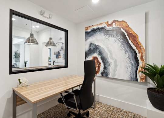 A black chair sits in front of a wooden desk in a room with a large painting on the wall.