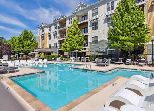 A large swimming pool surrounded by lounge chairs and umbrellas in front of a multi-story apartment building.