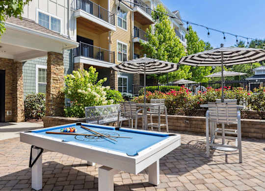 A pool table is surrounded by chairs and umbrellas in a courtyard.