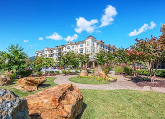 A large rock formation is in the foreground of a well-manicured lawn in front of a large building.