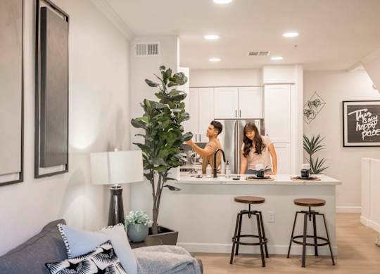 Two people are standing in a kitchen with a bar stool in front of them.