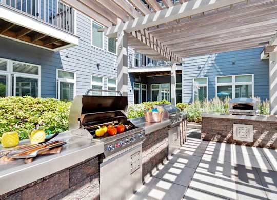 A BBQ grill is on a patio with a stone wall and a building in the background.