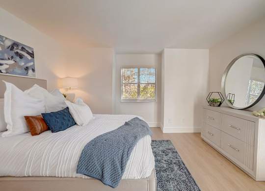 A bedroom with a large bed, a round mirror, and a window with a view of trees.