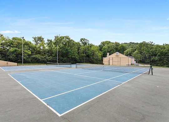 A tennis court with a blue surface and white lines, surrounded by trees and a building in the background.