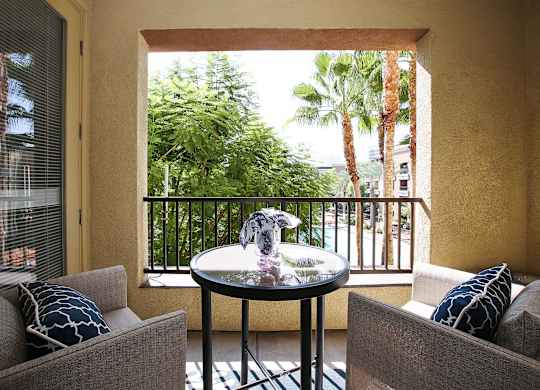 A balcony with a table and two chairs overlooking a lush green area at The Kitt at Warner Center Apartments, California
