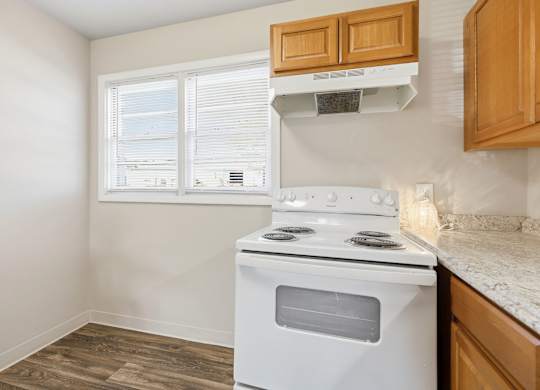 A white stove in a kitchen with wooden cabinets and a window with blinds.