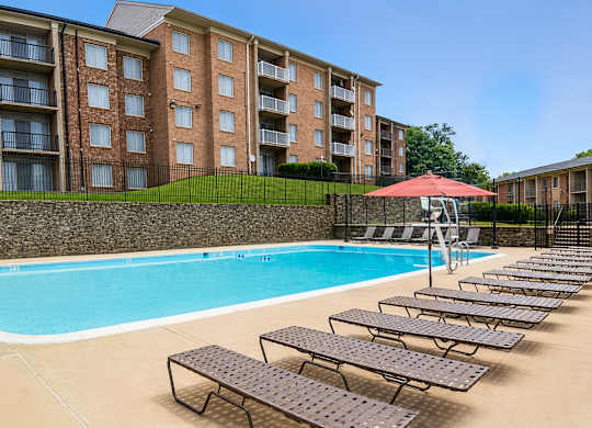 A swimming pool with sun loungers and a red umbrella in front of a brick building.