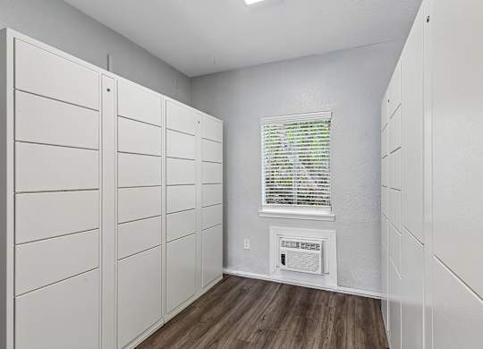 A room with white walls and wooden floors, featuring a row of white cupboards and a window with blinds.