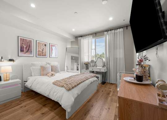 A bedroom with a large bed, a nightstand with a lamp, a dresser with a mirror, and a window with curtains.