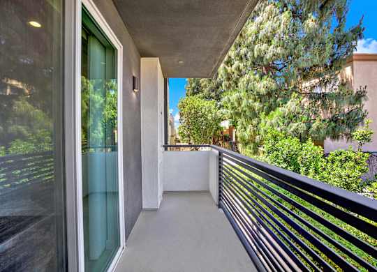 A balcony with a black railing and a glass door leading to a lush green outdoor area.