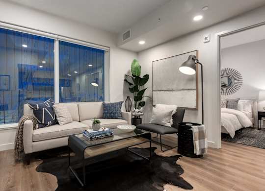 A modern living room with a large window, a sofa, a coffee table, and a bed in the background.