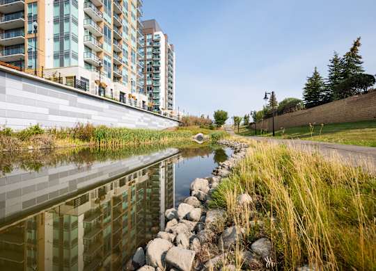 A river runs through a grassy area next to a concrete wall and apartment buildings.