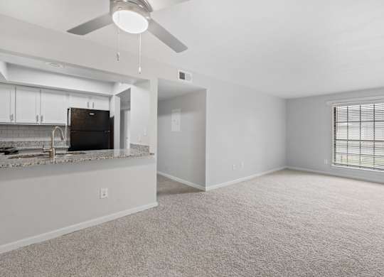 A spacious living room with a ceiling fan and a kitchen area in the background.