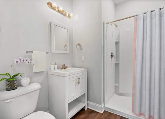 A white bathroom with a white tub, a white cabinet, a white toilet, a white sink, a white mirror, a white towel, a white vase with purple flowers, a white tray with a soap dish