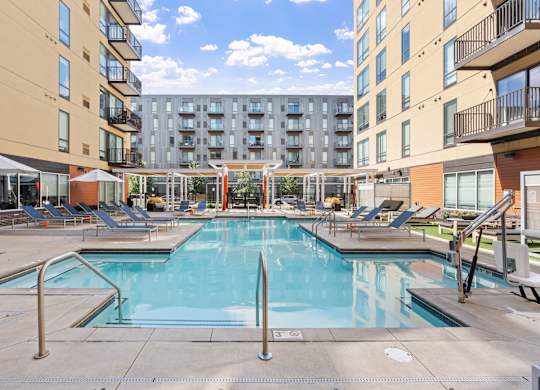 A large swimming pool surrounded by lounge chairs and umbrellas in a sunny courtyard.
