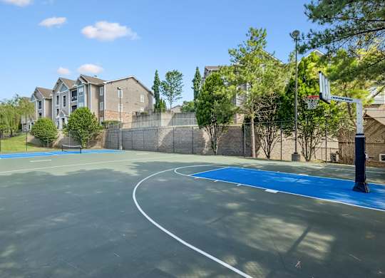 A basketball court with a blue surface and white lines, surrounded by a fence and houses.