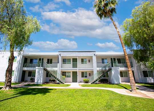 A white building with a balcony on the second floor is surrounded by green grass and trees.