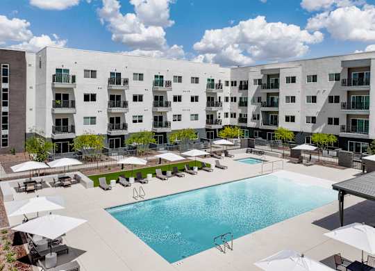 A large swimming pool surrounded by lounge chairs and umbrellas in front of a white apartment building.