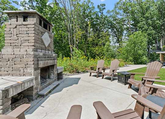 A stone fireplace is in the middle of a patio with wooden chairs around it.