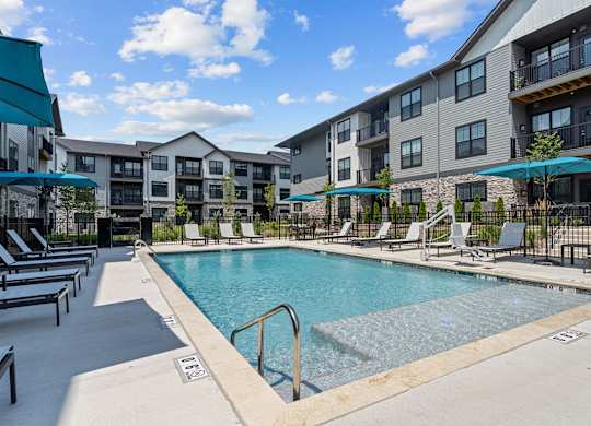 A swimming pool surrounded by lounge chairs and umbrellas in front of apartment buildings.
