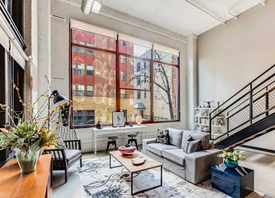 A living room with a grey couch, a coffee table, a staircase, and a large window.