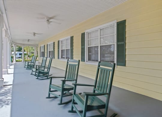 a row of green rocking chairs sit on the porch of a yellow house with green shuttersat Arbor Oaks at Greenacres, Greenacres, FL 33467