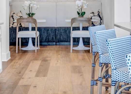 A white chair with blue patterned cushions is in the foreground of a room with a wooden floor and a white wall with a decorative mural.