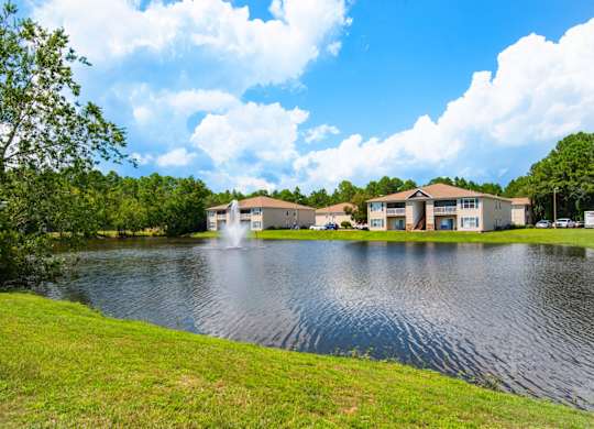 A serene pond with fountain display surrounded by greenery and apartment buildings at Crystal Lake in Pensacola, Florida.