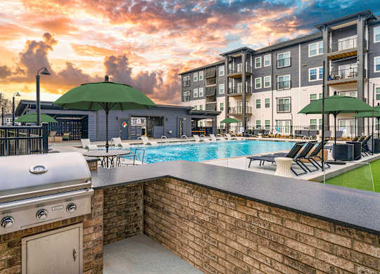 A sunset view of a pool area with a barbecue grill and lounge chairs.