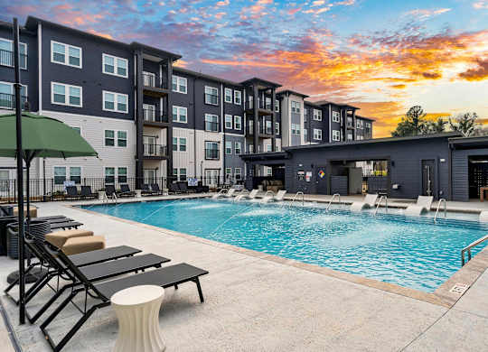 A swimming pool surrounded by lounge chairs and umbrellas in front of a modern apartment building.