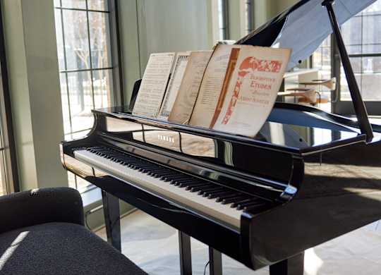 A grand piano with sheet music on top is placed in a room with large windows.