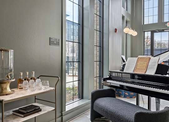 A room with a piano, a table with bottles and a book, and a window with a view of a building.