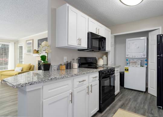 A galley-style kitchen with white cabinets, black appliances, granite countertops, and a washer and dryer in a laundry closet at Summerchase at Riverchase in Hoover, Alabama.