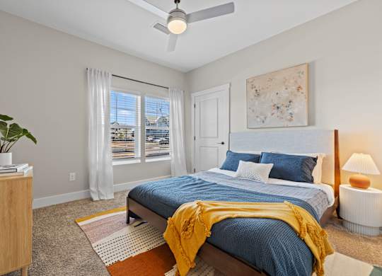 A bedroom with a bed, a ceiling fan, and a window with a view of buildings outside.