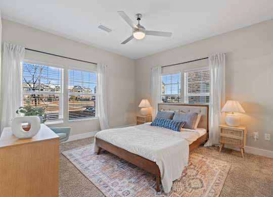 A bedroom with a large bed, a ceiling fan, and a view of the outdoors through the windows.
