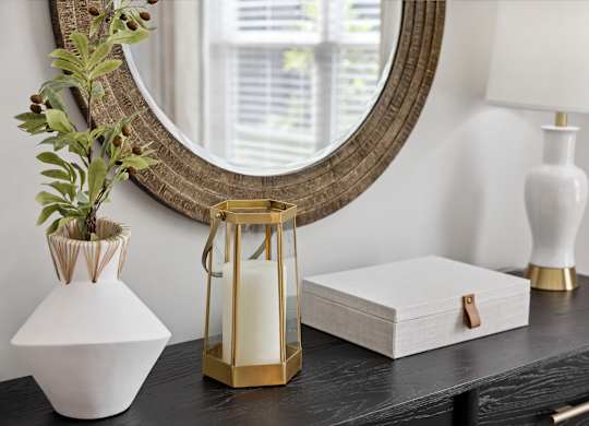 A white vase with a plant in it sits on a table next to a mirror and a white lamp.