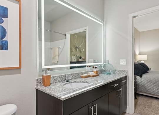 A bathroom with a sink, mirror, and a bed in the background.