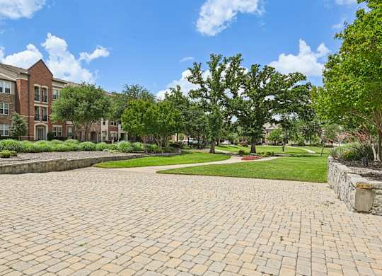 A brick pathway leads to a grassy area with trees and a building in the background.