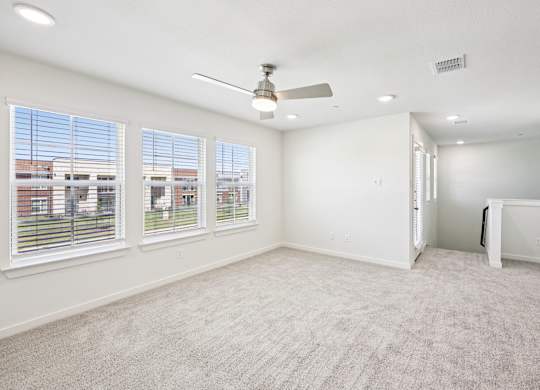 A room with a carpeted floor, a ceiling fan, and a view of a building outside the window.