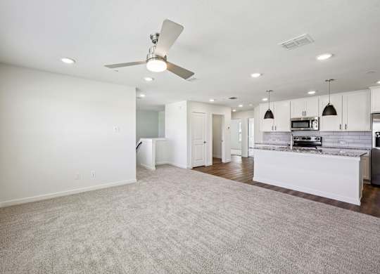 A spacious living room with a ceiling fan and a kitchen area in the background.