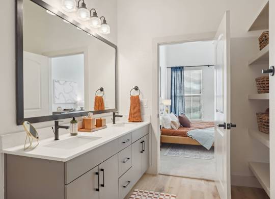 A bathroom with a vanity, mirror, and a bedroom in the background.