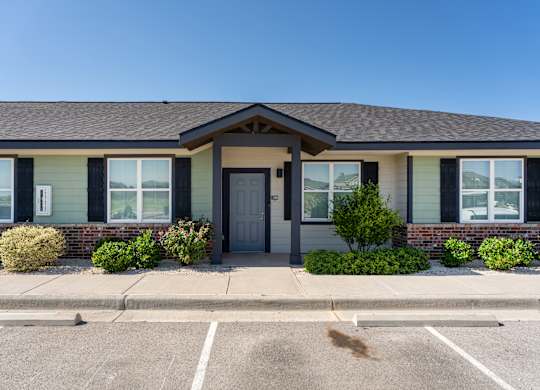 A building with a grey roof and a black door is surrounded by bushes.