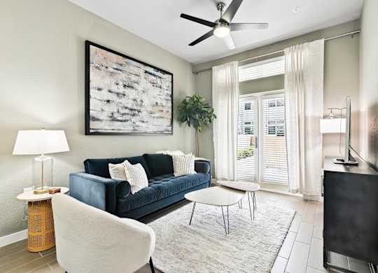 A living room with a blue couch, a white chair, a small table, a ceiling fan, and a painting on the wall.